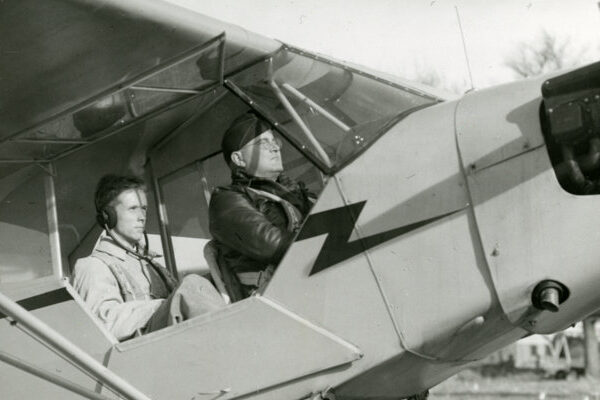 Black and white photograph with a student and his instructor, both dressed in flight gear, sitting in the cockpit of a small airplane.