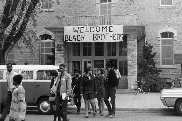 Black and white photograph with students walking away from a large stone building with a white banner hanging on front saying “Welcome Black Brothers” in large black letters.