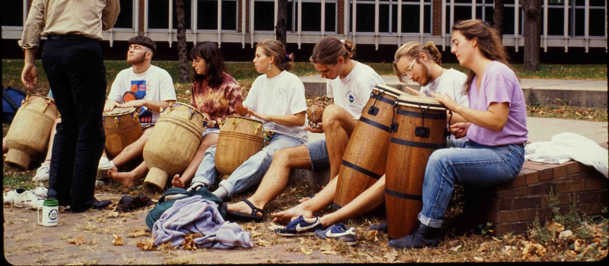 Surrounded by their jackets and shoes, seven students sit in front of a brick building and play bongo drums. Part of the photograph is blocked by a standing individual.